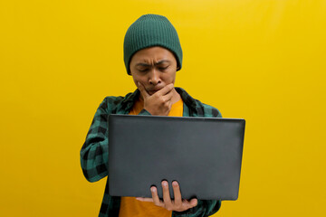 A young Asian freelance worker appears puzzled and lost in thought as he gazes at his laptop while...