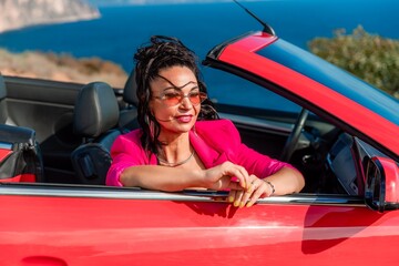 A woman in a pink jacket is sitting in a red convertible. She is wearing sunglasses and has her...