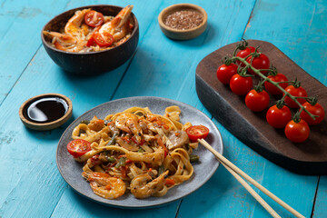shrimp udon pad thai sauce gray plate on a blue table next to cherry tomatoes soy sauce and...