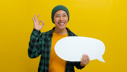 Excited Asian man in a beanie hat and casual shirt shows an OK gesture to the camera while holding a speech bubble with copy space. Isolated on a yellow background