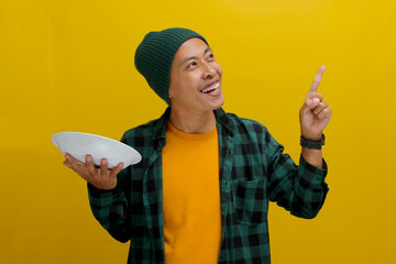 An excited young Asian man, dressed in a beanie hat and casual shirt, points at an empty space...