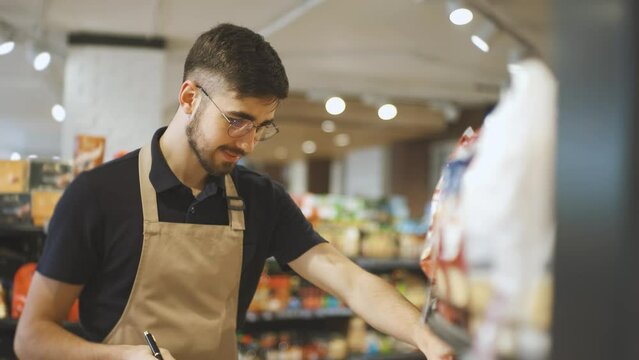 Man in a black t-shirt and a apron walks along the a supermarket and takes inventory using paper and a tablet