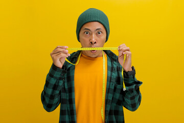 An enthusiastic young Asian man, clad in a beanie hat and casual shirt, gazes at a measuring tape with an expression of surprise or excitement, mouth slightly agape, against a yellow background
