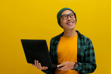 Confident and cheerful Asian businessman, dressed in beanie hat and casual shirt, holds a laptop...