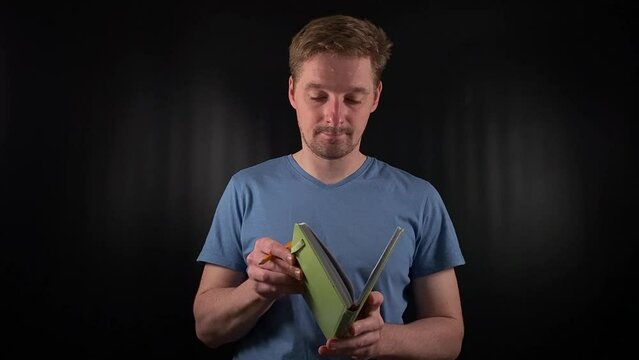 A portrait of a handsome young white man holding a notebook, opening it, jotting down notes, making entries in a planner on a black background. Planning and record-keeping in a purposeful lifestyle