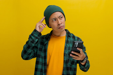 Pensive Asian man, dressed in a beanie hat and casual shirt, holds a mobile phone, appearing...