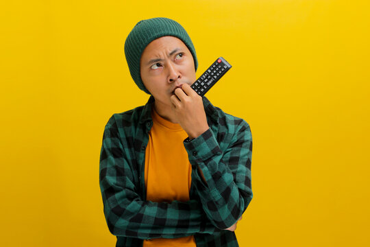 Pensive young Asian man, dressed in a beanie hat and casual shirt, appears puzzled while selecting a movie to watch, holding a TV remote at home, while standing against yellow background