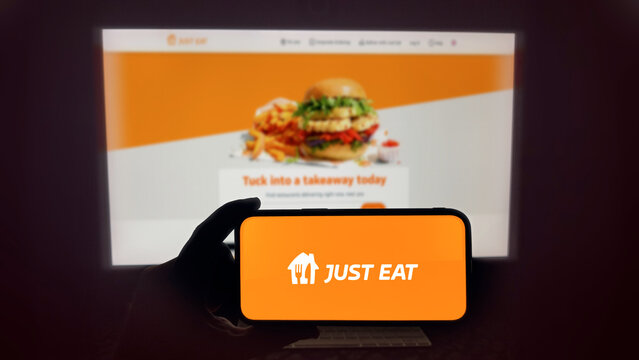 Just Eat food delivery app running on a modern mobile phone. The smartphone is being held in landscape mode, in the darkness. A PC monitor with the Just Eat website in the background.