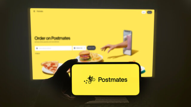 Postmates food delivery app running on a modern mobile phone. The smartphone is being held in landscape mode, in the darkness. A PC monitor with the Postmates website in the background.