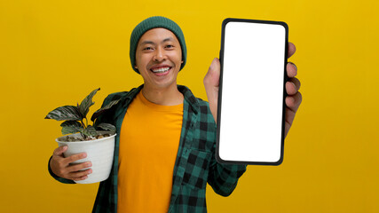 Cheerful Asian man showing his phone screen to the camera while holds a healthy Pin-stripe Calathea...