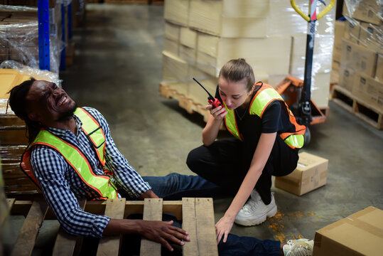 African American worker having accident and injured leg while working in warehouse while his women colleague is calling the rescue team. safety workplace and emergency situation support concept.