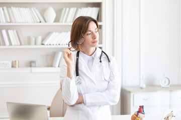 Thoughtful female doctor cardiologist in white medical gown standing in front of desk in the modern...