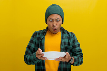 Surprised Asian man, dressed in beanie hat and casual shirt, holds an empty white plate in his...