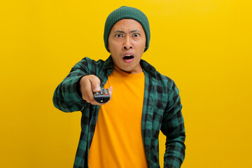 Surprised young Asian man, clad in a beanie hat and casual shirt, watches shocking news on TV while...