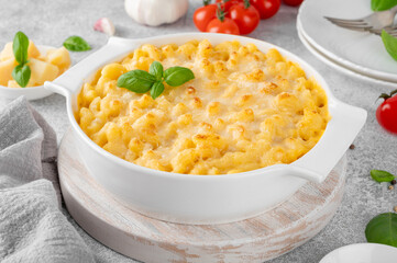 Mac and cheese in a white baking dish on a wooden board on a gray concrete background. American cuisine, comfort food. Copy space.