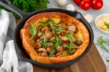 Savory Dutch baby pancake with cheese, fried mushrooms and arugula in cast iron pan on a concrete background. Vegetarian breakfast.