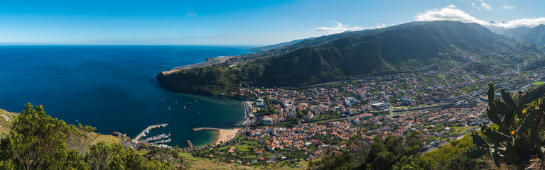 Panoramic aerial view of Machico bay with golden sand beach, palm trees from Pico do Facho viewpoint over the Machico valley, Airport in the background, Madeira, Portugal