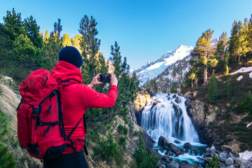 A man with a red jacket and backpack takes a picture of a waterfall with his mobile phone....