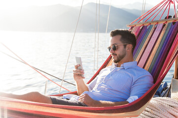 Man with mobile phone relaxing on yacht in hammock. Sea travel on sailboat. Traveler using...