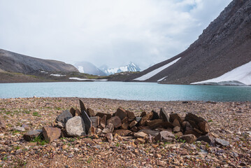 Naklejka premium Sharp pointed stones on background azure alpine lake against snow-covered few pointy peaks in low clouds. Ripples on blue water surface of mountain lake and three snowy peaked tops in low cloudiness.