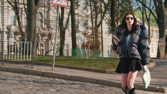 Astonishing high young lady cross the road with model-walk wearing sunglasses winter jacket short skirt and high black boots carrying laptop and her phone 4k video. Mooving in road in slow motion. 