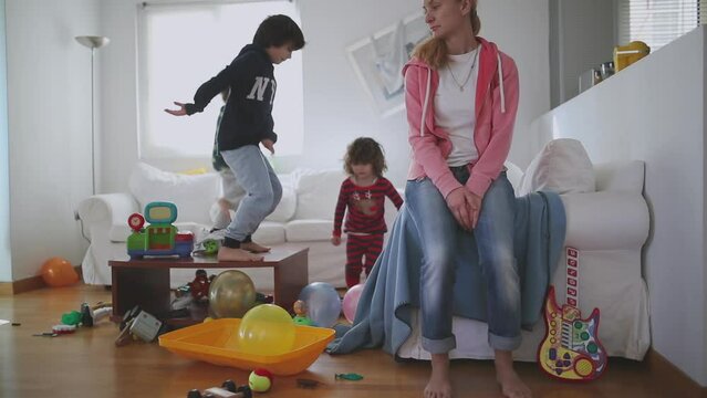 Exhausted mother sits with eyes closed and lying back on a couch, surrounded by her playful children and a disheveled living room strewn with toys, slow motion.
