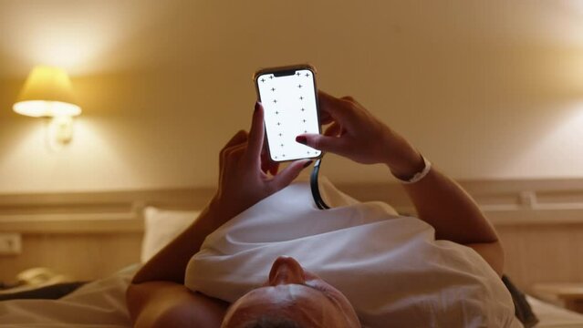 A Woman Lies on the Bed and Scrolls Through Her Smartphone Screen with a Mock-up of a White Screen. Evening in the Hotel Room, Concept of Ordering Food or Browsing the Menu.