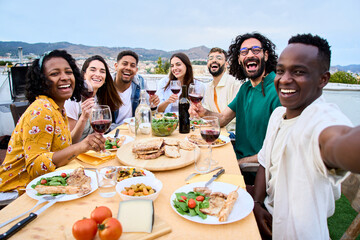 Young African smiling man taking selfie to joyful millennial people sitting at barbecue table....