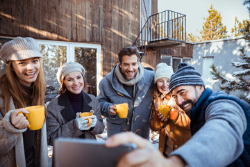 Group of friends enjoying winter drinks outdoors and taking a selfie in front of a cabin