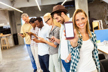 Smiling young woman showing smartphone with blank screen with diverse colleagues in background at...