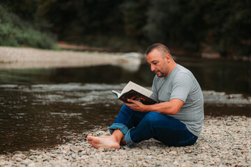 Younger man reading book by the river. Copy space