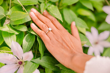 A tan skin woman has her hands over leaves and greenery in a garden. She wears a solitary diamond...