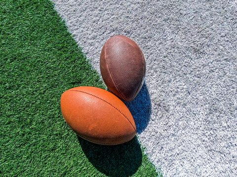 Two American Footballs on a Football Field. Two footballs lay on the ground with shadows. Slanted football field turf background in green and white. - Powered by Adobe