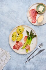Green boiled asparagus served with boiled potatoes, thinly sliced smoked pork and sauce on a round clay plate on a gray concrete background. Asparagus recipes.