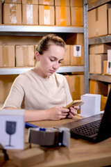 Fototapeta premium Young entrepreneur small business checking internet orders on smartphone and preparing boxed items for delivery to customers