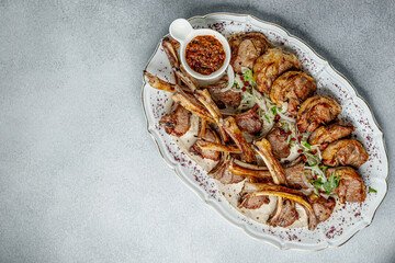The photo shows roasted ribs neatly laid out on a white platter with ornaments. The ribs are well fried and garnished with herbs. The background of the image is a textured gray table, which contrasts 