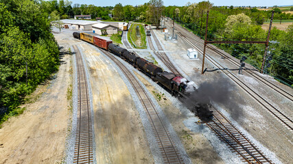 Aerial vintage steam train moves through industrial rail yard, emitting steam as it passes warehouses alongside other rail cars