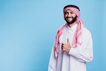 Muslim man dressed in traditional clothes showing thumb up while standing with cheerful expression...