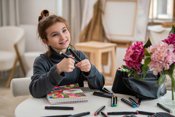 A young girl is sitting at a table with a notebook and a bag of markers.
