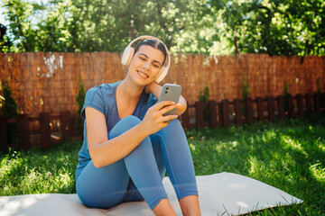 One young caucasian woman is sitting on yoga mat with wireless headphones and mobile phone preparing for training	
