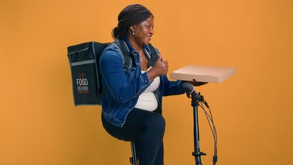 For prompt and dependable delivery service female cyclist on bicycle transports package from restaurant. African american courier with takeout box and a thumbs up ensures eco-friendly food delivery.