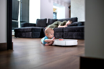 Cute young woman is sleeping on sofa in living room while little child is playing with robot vacuum...
