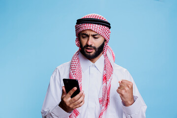 Excited muslim man wearing islamic traditional clothes reading news on social media using...