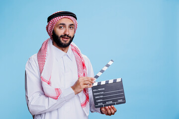 Muslim man dressed in thobe and headscarf clapping movie slate, showing film scene action studio...