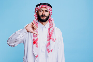 Muslim man dressed in traditional arabian clothes showing thumb down studio portrait. Arab in thobe and ghutra showcasing disagreement and looking at camera with serious expression