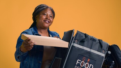 Youthful black woman courier taking out pizza box from food delivery bag. Efficiency meets...