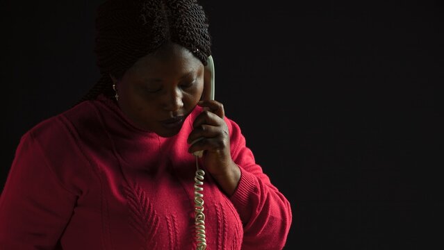 Side-view portrait of black woman with pink sweater standing with handset wired phone unsatisfied by the talk. Young african american female having a serious conversation on landline telephone.