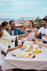 Vertical. Group of happy young people toasting red wine and celebrating meal on rooftop....