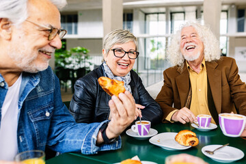 Happy elderly people enjoying brunch. Group of senior retired friends having fun drinking hot coffee while social gathering sitting on bar table. Retirement lifestyle concept.