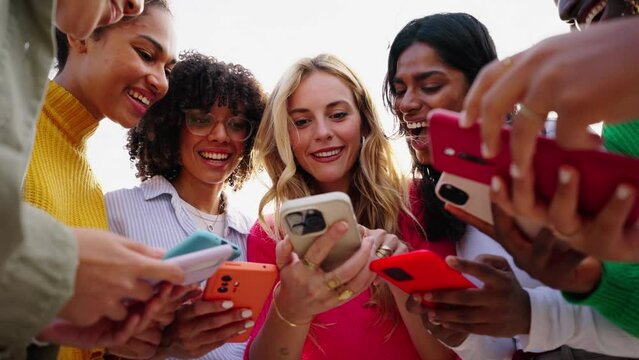 Low angle. Girls time. Group of multiracial young only women smiling gathered in circle using mobile phones. Cheerful females looking cells concentrated. Addiction to technology of gen Z